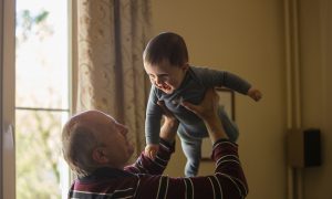 man wearing maroon, white, and blue stripe long-sleeved shirt lifting up baby wearing gray onesie