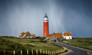 asphalt road between grass field going to brown lighthouse beside houses under cloudy sky