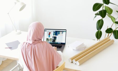 A woman wearing a hijab is working on her laptop at a modern desk with indoor plants and natural light.