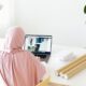 A woman wearing a hijab is working on her laptop at a modern desk with indoor plants and natural light.